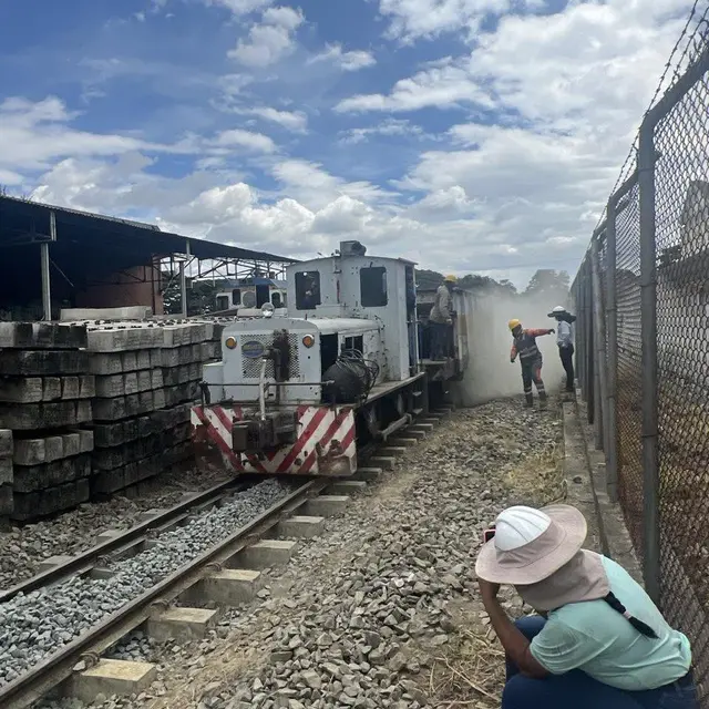 Locomotora de maniobras junto a pilas de traviesas de concreto, con una trabajadora en primer plano